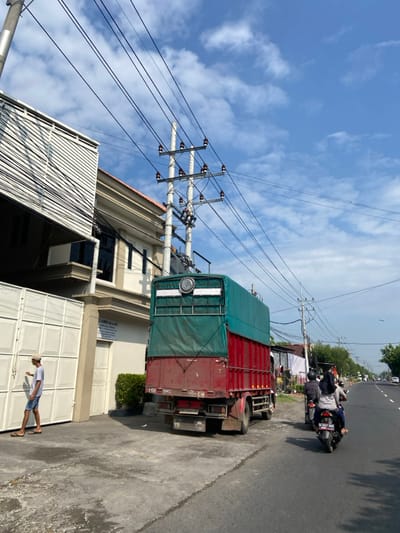 The image shows a street scene in Kecamatan Tanggulangin, Indonesia (latitude: -7.497, longitude: 112.685). A large, red and green truck is parked on the side of the road, facing forward. The truck has a tarp covering its cargo bed and is partially visible through the tarp. A man is standing on the sidewalk near a closed white gate, looking to the left. He is wearing a light blue shirt and shorts. On the other side of the truck, two men are riding motorcycles. The man on the front motorcycle is wearing a black helmet and a white shirt. The man on the back motorcycle is wearing a black helmet and a dark shirt. Both motorcyclists are wearing sandals. The motorcycles are facing forward and are in the middle of the street. On the opposite side of the road, a man is walking away from the camera. Behind the parked truck, there is a two-story building with white walls and a white corrugated metal roof. Power lines and power poles are strung across the street.  The day is sunny, with a blue sky and some white clouds. The road surface is paved and there is a sidewalk on the right side of the road. The image appears to be taken from a pedestrian’s perspective.