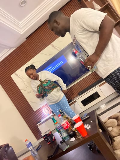Two individuals are seen in a modern indoor setting, likely a living area in Adenta Municipality, Ghana, engaged in a casual gathering. On the left, a male figure wearing a white t-shirt holds a clear spirit bottle with a red cap, appearing to pour its contents. He has short dark hair and is looking towards the right. On the right, a female figure is seated or crouched, wearing a white long-sleeved top, blue polka-dotted pajama bottoms, and a vibrant patterned scarf. She has dark braided hair and is looking forward.
In the center of the room, a large flat-screen television displays a "no signal" message with troubleshooting text: "Connect your Antenna cable is connected properly. Chanosi and perfum Auto Channel Scan". Below the TV is a white marble-patterned surface. To the right, a dark brown table is laden with various beverages, including multiple bottles of spirits, a Coca-Cola bottle, and water bottles. Two bright red plastic cups and other miscellaneous items like crumpled paper and electronic accessories are also scattered on the table. The decor features textured brown slatted wall panels and a white ceiling with recessed lighting, contributing to the contemporary feel of the space. The scene suggests a relaxed social event, likely taking place in the evening or night.