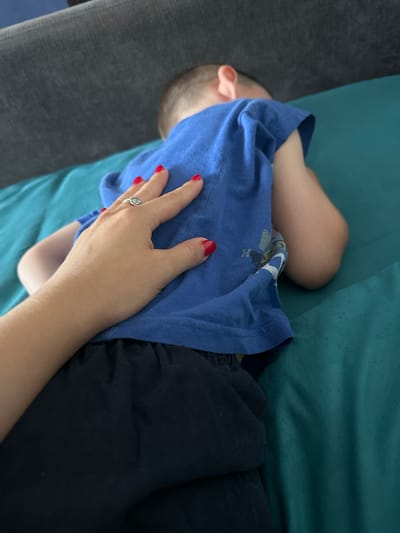 A close-up shot shows a hand with bright red nail polish resting on the back of a child wearing a blue t-shirt. The child is lying on their side on a turquoise surface, facing away from the camera. A grey sofa is visible in the upper left corner of the image. The hand wears a ring with a grey stone. The scene appears to be indoors, possibly in a living room or on a couch. There is no indication of the location within the city of Novoaltaysk, Russia.
