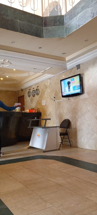 A hotel lobby features a dark front desk, three clocks on the wall, and a TV playing news. A small, silver event table stands beside a black folding chair. The floor is tiled with beige and green marble. The ceiling is layered with crown molding and ornate details. A wrought iron balcony is visible above.