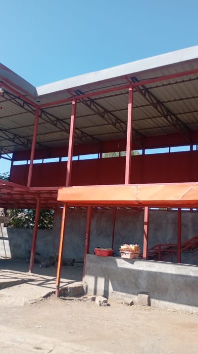 An open-air structure stands with a corrugated metal roof supported by red metal beams. The second story has a red wall with rectangular openings. A concrete base supports the lower level, where baskets of goods rest on the ledge. The ground is dirt, and a clear blue sky is above.