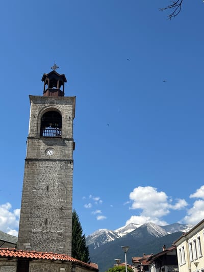The image shows the historic bell tower of the Holy Trinity Church in Bansko, Bulgaria. The tower is made of stone and has a clock face. Above the clock, there is an arched opening with bells inside. The tower is topped by a wooden structure with a cross. In the background, there are snow-capped mountains and a clear blue sky with some clouds. Several birds are visible in the sky. The area around the tower appears to be a town with buildings and red-tiled roofs.