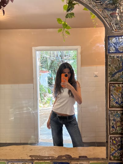 The image shows a young woman standing in front of a mirror. She is wearing a white tank top and blue jeans. She is holding a smartphone with an orange case in front of her face, covering her eyes. The background is a bathroom with a white tiled wall and a doorway leading to an outside area with greenery. The woman appears to be taking a selfie. The mirror frame is decorated with ornate tiles. The scene takes place in the city of Ra'anana, Israel, specifically at the coordinates 32.173 latitude, 34.872 longitude.