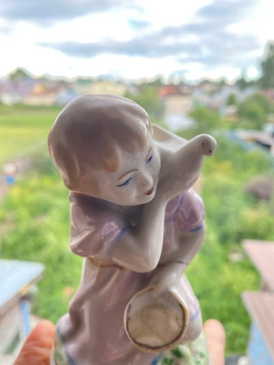 A close-up, side-angle shot shows a porcelain figurine of two figures, possibly children, embracing. The figurine is predominantly white with hints of pink and blue. The face of one figure, closer to the camera, is detailed with closed blue eyes and pink cheeks. The figures are positioned in a way that suggests affection or rest. The background is softly blurred, showing a landscape with green foliage and hints of buildings, suggesting an outdoor setting during daytime. The lighting appears natural and bright, indicating it might be midday. The image is captured with a shallow depth of field, focusing on the figurine while blurring the surroundings. The context provided indicates the location is Zelenodol'sk, Russia. There is no visible text in the image.