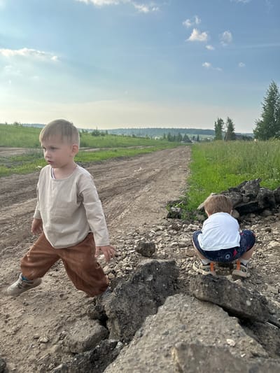 Two young boys are in a rural outdoor environment, likely in the vicinity of Estachi, Russia. The scene depicts a dirt road that stretches into the distance, flanked by grassy fields and scattered trees. In the foreground, a dirt pile composed of rocks and what appears to be broken asphalt is visible.
The boy on the left is facing towards the right of the frame, mid-stride. He has light blonde hair and is wearing a loose-fitting, long-sleeved beige shirt and brown pants. His right hand is extended outwards.
The boy on the right is positioned in a crouched stance, facing away from the camera. He has blonde hair and is wearing a white t-shirt and patterned shorts. His feet are clad in sneakers with orange accents.
The sky above is partly cloudy with a clear blue expanse. The overall lighting suggests it is daytime, possibly late afternoon or early evening given the warm tones. The environment appears undeveloped and somewhat rugged.