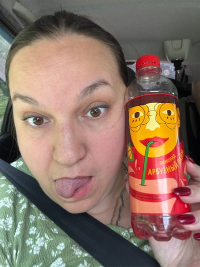 A woman is pictured indoors, holding a bottle of lemonade. She is in the driver's seat of a car, indicated by the seatbelt and dashboard visible. The woman has brown eyes and is sticking her tongue out, giving a playful expression. The lemonade bottle has a cartoon drawing of a face with glasses and a straw. The label on the bottle is in Russian and reads "ЛИМОНАД АРБУЗНЫЙ" which translates to "Watermelon Lemonade". The woman is wearing a green floral print shirt. The background is blurred, but it appears to be a natural landscape. The image was taken on August 2, 2023.
