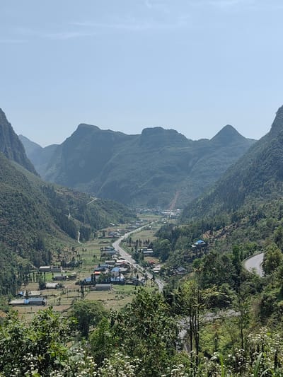 The image depicts a scenic view of Phố Bảng Commune, Vietnam. The foreground features lush greenery with white wildflowers and shrubs. A winding road cuts through the hillside. In the valley below, there is a village with houses and farmland. A road runs through the center of the village. The background consists of mountains covered in dense forest, stretching into the horizon under a clear sky.