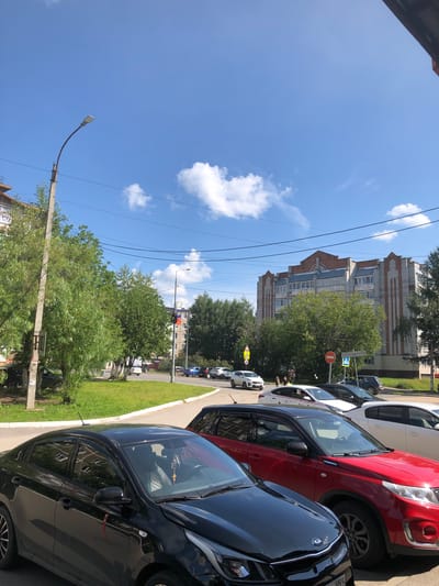 The image captures a street scene in Solikamsk, Russia, on a sunny day with a bright blue sky and scattered white clouds. In the foreground, a black sedan is parked, followed by a red SUV and a white sedan. Several other vehicles are visible further down the street, including a blue car. Trees line the sidewalk and a grassy area separates the road from a multi-story brick building in the background. A lamppost stands on the left side of the frame, and traffic signs are visible on the right. A flag, possibly a Russian flag, is attached to a lamppost.