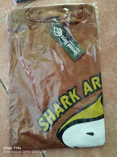 A brown t-shirt with the words "SHARK ARMY" written in bold, yellow lettering is lying flat on a brown surface. The t-shirt is wrapped in clear plastic and a white rectangular sticker is attached to the upper right corner of the t-shirt with the words "Second Boys - Original Kidswear" written in black. The location of the image is Anjatan, Indonesia, specifically at latitude -6.419 and longitude 107.978.