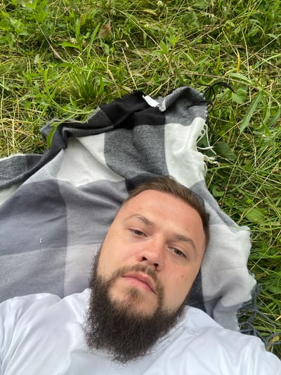 A man with a full beard and brown eyes is lying on a checkered blanket outdoors. The blanket is gray and white with black accents. He is wearing a white shirt. The blanket is placed on a grassy area with some weeds and plants. The image is taken from a high angle, looking down at the man. The lighting suggests it is daytime, and the greenery indicates a possibly warm or mild weather. The man's expression is neutral. There are no visible text elements in the image.