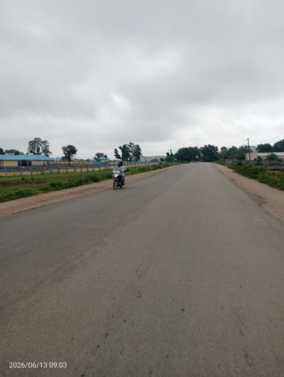 This is a photograph taken on a road in Suleja, Nigeria. The sky is overcast with grey clouds. The road itself is paved and appears to be a two-lane highway with no visible lane markings. The road stretches into the distance, with the perspective suggesting the photo was taken from a moving vehicle or while standing on the roadside.
On the left side of the road, there is a yellow and blue fence bordering a grassy area. Beyond the fence, there are several low-rise buildings with blue roofs, appearing to be part of an institutional complex. Trees are scattered around this area.
On the right side of the road, the terrain is also bordered by a dirt path and overgrown vegetation. In the distance, more trees and some buildings are visible, including what looks like a power pole with cables.
A motorcycle is the only visible vehicle on the road in this frame. It is traveling away from the camera, carrying two individuals. The rider is wearing a white shirt and a dark cap. The passenger is also wearing a light-colored shirt.
The date and time stamp in the lower left corner indicates "2026/06/13 09:03".