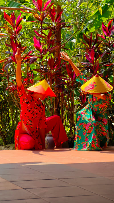 This vivid image captures two individuals, likely women, performing a graceful dance or pose on a tiled patio in Thông Nguyên Commune, Vietnam. They are dressed in traditional Vietnamese attire, characterized by vibrant floral patterns and iconic conical hats (Nón Lá), set against a backdrop of lush tropical foliage.

The person on the left wears a bright red long-sleeved tunic with intricate red floral designs, complemented by solid red loose trousers. Their conical hat is predominantly red, featuring a sparkling gold section at its peak. This individual is depicted in a kneeling or crouched position, with one arm elegantly raised and fingers gracefully splayed.

To the right, the second person dons a flowing green garment adorned with striking red floral patterns. Their yellow conical hat is also decorated with similar red floral motifs. This individual is in a lower, seated or deeply crouched pose, with both arms raised in a delicate, expressive gesture.

The scene is bathed in bright daylight, evident from the strong shadows cast on the terracotta-colored tiled floor, indicating a sunny day, possibly around midday. The surrounding environment consists of dense green and red-leafed plants, adding to the tropical ambiance. The overall composition highlights the beauty of Vietnamese culture and tradition within a vibrant natural setting.