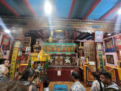 The image shows the interior of a Buddhist temple, likely the Manu Temple, located in the city of Manali, India. A large, golden Buddha statue is prominently displayed in the center, seated on a decorated altar. The statue is surrounded by various religious artifacts, including smaller Buddha figures, paintings, prayer flags, and shelves filled with religious texts. The temple's ceiling features exposed wooden beams painted in red and blue. Several individuals are present in the foreground, observing the central Buddha statue. The lighting is bright, with sunlight streaming in from the right side of the image, creating lens flare effects. The walls and structures of the temple are adorned with intricate Tibetan Buddhist art. A sign in Hindi and English, reading "DO NOT TOUCH ANYTHING," is visible near the altar. The atmosphere appears serene and reverent, indicative of a place of worship.