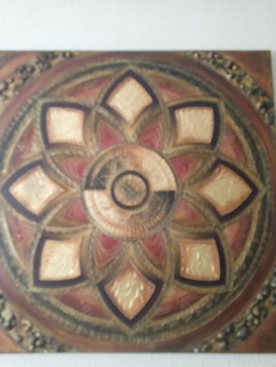 The image displays a close-up, blurred view of a circular, mandala-like artwork. The artwork features intricate geometric patterns with a color palette dominated by earthy tones, including browns, reds, and gold. The central area of the mandala contains concentric circles with contrasting dark and light segments. Surrounding the center are petal-like shapes and diamond-shaped insets filled with a textured, golden material. The outer edge of the artwork appears to be a textured border with small, dark, pebble-like elements. The artwork is mounted on a wall. The location of this artwork in Ильинское-Усово, Russia, is not discernible from the provided image.