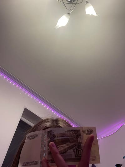 A person holds a Russian 10 ruble banknote in front of their face, obscuring their features. The banknote displays the number "10" in several locations and has Cyrillic text that reads "ДЕСЯТЬ" (ten). The person's hair is visible, and a portion of their hand with painted nails is also present, with a finger extended towards the banknote. The background is a room with a white ceiling featuring a modern light fixture with three tulip-shaped glass shades. Along the top edge of the wall, a string of purple LED lights is visible. The overall lighting suggests an indoor setting at night. The image does not contain any visual information that would allow for the determination of its location within the city of Romanovka, Russia.