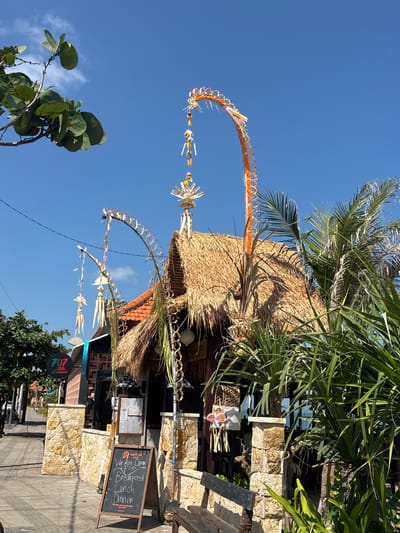 This is a medium shot, eye-level, outdoor, daytime photograph taken on a sunny day in the city of North Kuta, Indonesia. The foreground features a sidewalk and a building with a thatched roof, adorned with traditional Balinese decorations that appear to be made of woven palm leaves and flowers. A blackboard sign on the sidewalk advertises "We Are Open for Breakfast, Lunch, Dinner" and "The Wine that Speaks Bali Est. 1994." The building is constructed with stone pillars and dark wood, and tropical plants, including palm trees, surround it. In the background, the sky is clear blue with a few wispy clouds. A street with other buildings and trees is visible to the left, and a glimpse of the ocean can be seen beyond the building. There are no people visible in the image.