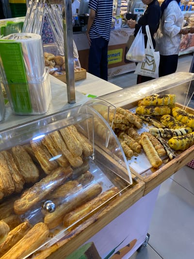 A street-level view shows a bakery display cart filled with assorted pastries, situated in a well-lit indoor market or supermarket. On the left, rolls of transparent plastic bags with green patterned edges are stacked. The cart itself has wooden trim and a light purple base. The pastries are arranged in clear plastic containers and wooden trays. Some pastries are long and golden brown, sprinkled with what appears to be cheese or sugar, while others are filled with chocolate and topped with chocolate shavings. In the background, a woman in a black top and hijab stands with shopping bags, holding a phone. Another woman in a white patterned top is also visible. A person wearing a striped shirt is to the left of the frame. The environment appears to be a busy commercial space, possibly a supermarket or a market hall in Madiun, Indonesia, given the signage that mentions "ermart" and "and more." The overall scene depicts a food retail setting with a focus on baked goods.