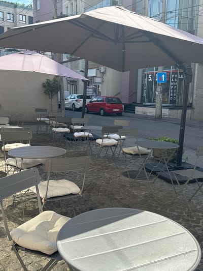 The image depicts an outdoor seating area of a cafe on a paved street in Vračar Municipality, Serbia. Several gray metal folding tables and chairs, some with light-colored cushions, are arranged on a cobblestone surface. Large umbrellas, one light pink and another a muted brown, provide shade. In the background, a red car is parked, and other vehicles are visible on the street. Buildings line the street, some with glass facades and signs, including one with the text "Always Be Ready" and another with numbers and letters. The environment suggests a daytime scene with ample sunlight casting shadows.