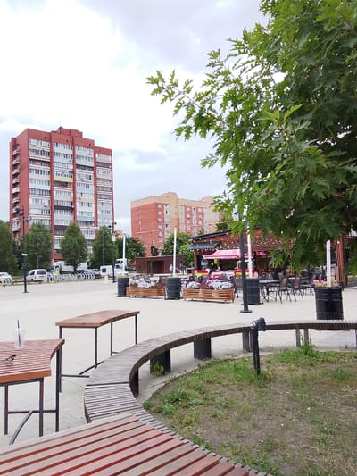 This outdoor scene in Orekhovo-Zuyevo, Russia, depicts a public space with several key elements. In the background, two tall residential buildings, one brick-red with numerous balconies and windows, and another to its right with a lighter brick facade, dominate the skyline. Between them, a smaller, modern, wooden-structured cafe or restaurant with outdoor seating is visible, featuring a red umbrella and string lights.
In the foreground, several pieces of outdoor furniture are arranged on a paved area. These include long, slatted wooden benches, some curved and others straight, and wooden tables with black metal legs. One table has a clear plastic cup with a straw on it. A small bird is perched on this table. Large black cylindrical planters are placed near the cafe and benches. The area appears to be a pedestrian zone, with a bike rack and parked vehicles, including vans and cars, visible in the middle ground.
The weather is overcast, suggesting it might be a cloudy day. The time of day is indeterminate due to the even light. The overall atmosphere is that of a public urban space, perhaps a park or a town square, designed for relaxation and social gathering, with elements of both residential and commercial life present. There are no explicit signs of people actively engaged in activities, but the presence of tables and benches implies potential use. The green foliage of a large tree in the foreground adds a natural element to the urban setting.