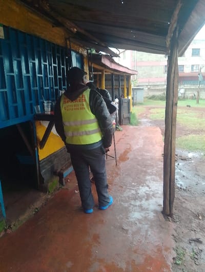 The image shows a man standing in front of a shop in Gitaru, Kenya. The man is wearing a yellow vest with a red inscription, "CYCLE SPARES," with two phone numbers, "0715 353 333" and "0712 888 888." He is also wearing blue shoes and black pants. The shop has a blue door and a sign above it. There is another person partially visible in the image. The scene is partially covered by an awning, and the ground is wet with water. The environment appears to be rural with a dirt ground and a small building in the background.