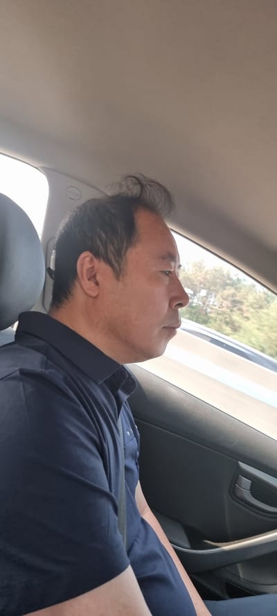 An Asian man is seen from the chest up, resting in the passenger seat of a car. He wears a dark blue shirt and has a seatbelt across his chest. He is looking up and slightly toward the window. The vehicle is moving. The location is Ucheon-myeon, South Korea; this is inferred from the foliage visible through the car window.