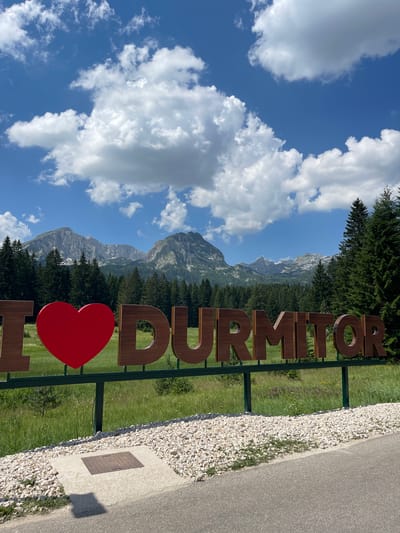 The image shows a scenic view of Durmitor National Park in Pitomine, Montenegro. In the foreground, large wooden letters spell out "I LOVE DÚRMITOR," with a prominent red heart replacing the word "LOVE." The letters are mounted on a green metal frame. Behind the sign, a lush green meadow is dotted with wildflowers. A dense forest of evergreen trees stretches across the middle ground, leading to a majestic mountain range in the background. The mountains are rugged and rocky, with some patches of snow visible on their peaks. The sky is a vibrant blue, filled with scattered white cumulus clouds. The lower right corner of the image shows a portion of an asphalt road and a gravel path with a manhole cover set in concrete.