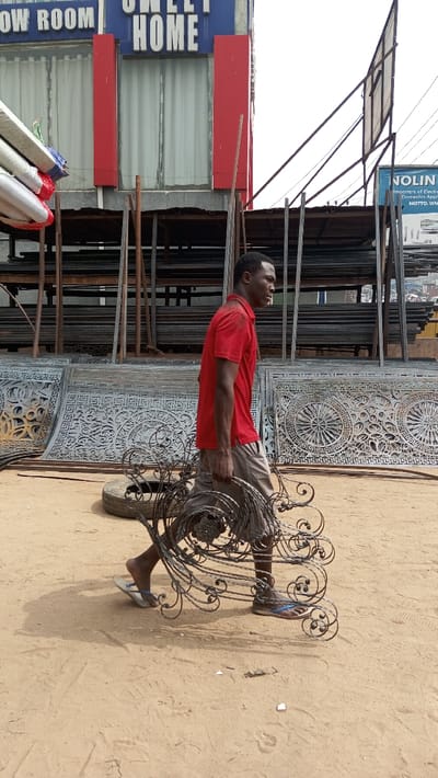 An adult male is depicted walking from left to right across unpaved, sandy ground in Awkuzu, Nigeria. He wears a red polo shirt, light-colored shorts, and blue and white flip-flops, carrying several dark, intricately scrolled metal decorative pieces in his right hand. His shadow and that of the objects are cast on the ground. The immediate background features stacked ornate metal panels with geometric and floral patterns, alongside a discarded tire and a large metal rack holding numerous horizontal metal bars. In the upper background, a building with a blue facade and red vertical accents displays "OW ROOM" and "SWEET HOME" in white lettering, with white curtains visible in its windows. To the right, a metal billboard structure stands, showing a partial sign with text including "JOLIN" and "IMPORTERS of Electronics, Domestics Appliances." The scene is outdoors under bright, daylight conditions.