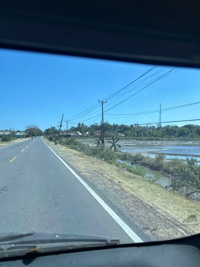 The image captures a view from inside a vehicle on a paved road in Jeneponto, Indonesia. The road is bordered by dry grass and vegetation. To the right of the road, there are salt fields, which appear to be shallow ponds with water. In the distance, a tower stands against a clear blue sky. There are also several power lines and utility poles visible along the road and in the background. A few motorcycles are parked along the side of the road, and a couple of people can be seen riding motorcycles in the distance on the left side of the road. Some houses and trees are scattered throughout the landscape. The overall scene depicts a rural or semi-rural setting under bright daylight.