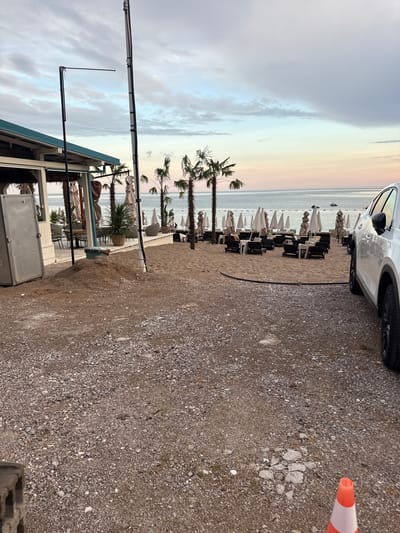 The image depicts a beach scene at Kamenovo, Montenegro, likely in the early morning or late afternoon given the soft, pastel hues of the sky. The foreground is dominated by a gravelly area with a white car parked on the right side, partially visible. A bright orange and white traffic cone is also present in this area.
The middle ground reveals a sandy beach lined with rows of lounge chairs and umbrellas, suggesting a resort or public beach area. Palm trees are scattered along the beach, adding to the tropical atmosphere. A small building or kiosk with a blue roof is situated on the left side of the beach, with some furniture and umbrellas visible around it. The sea stretches out to the horizon, meeting a sky filled with clouds painted in gentle shades of pink, orange, and blue.
The scene is quiet and appears to be either before or after a busy period, with no people clearly visible in the rows of chairs. The overall mood is serene and peaceful, with the soft lighting contributing to a tranquil ambiance. There is no visible text in the image.