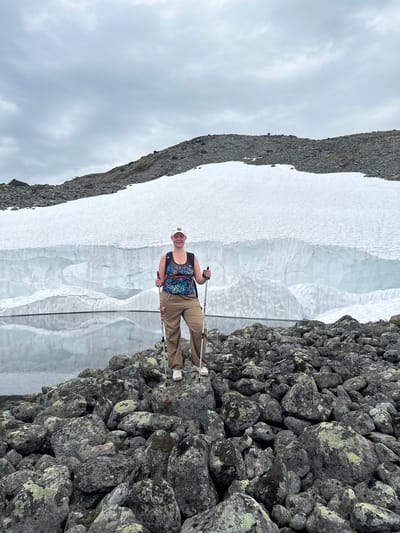 In Monchegorsk, Russia, an adult female individual stands facing the camera on an uneven field of dark grey rocks, many covered with green and yellow lichen. She is positioned in the lower-middle of the frame, holding a white and red hiking pole in each hand and smiling. She wears a white baseball cap, a patterned tank top primarily in blue, green, and black, a black mesh underlayer, light brown cargo pants, and white sneakers. A black backpack is visible on her back. Behind the individual, a calm body of water reflects a large, distinct white snow and ice wall that spans the mid-ground. The wall displays varied textures and layered formations. Above the snow and ice wall, a dark grey, rocky mountain slope with some snow patches rises towards an overcast, light grey sky.