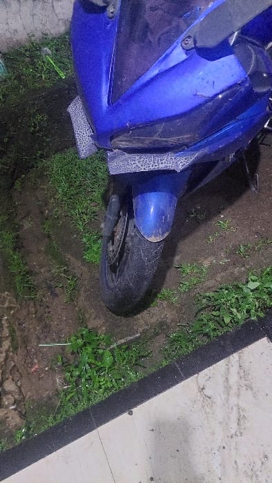 A blue motorcycle is parked on a dirt patch with green vegetation. The motorcycle's front tire is visible and the front fender is dirty. The motorcycle is partially obscured by green foliage. The scene is located in Kecamatan Babakan Madang, Indonesia, at the coordinates -6.579 latitude and 106.886 longitude. There are no people in the scene. The ground is covered in a thin layer of dirt and the surrounding vegetation is lush and green.