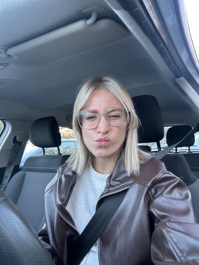 A blonde woman with glasses sits in a car, wearing a brown leather jacket and a white textured sweater. She is angled towards the camera, pursing her lips slightly. A black seatbelt is visible across her chest. The interior of the car shows a gray headliner, rearview mirror, and black headrests. Outside the window, glimpses of trees can be seen.