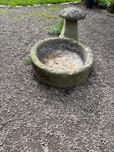 The image shows a stone bird bath in Pulverbatch, United Kingdom. The bird bath, made of weathered stone, features a rounded bowl atop a shorter, narrower stone pillar. The interior of the bowl is partially filled with light-colored material, possibly sand or sediment. The bird bath sits on a bed of small, gray gravel. To the left of the bird bath, green vegetation is visible along a border. The overall setting appears to be an outdoor area, possibly a garden or yard.