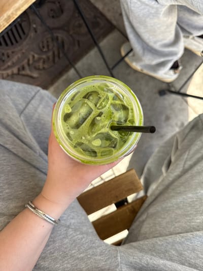 An overhead shot captures a person's left hand holding a clear plastic cup filled with an iced, foamy green beverage, likely matcha, containing several ice cubes and a black straw. The hand, with visible fingernails, rests over the person's lap, which is covered by light grey fabric, consistent with sweatpants. A silver-colored, twisted cable-design bracelet is worn on the left wrist. Below the hand and cup, brown wooden slats of a seat are visible. The background reveals a light-colored paved or tiled floor surface with a dark, ornate metal grate or manhole cover in the upper left. In the upper right background, the lower leg of another individual, clad in light grey fabric pants and light-colored footwear, is partially visible. The scene is illuminated by strong overhead light, casting discernible shadows. The location is Chula Vista, United States of America.