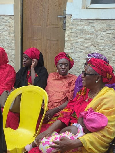 In Unguwar Rimi, Nigeria, a group of five individuals, primarily women, and one infant are seated outdoors against a building facade. On the left, a young woman, wearing a red patterned headwrap, a black outer garment over a red and yellow patterned top, sits on a bright yellow plastic chair. A grey handbag is visible beside her, and her right hand touches her ear. Adjacent, a woman in an orange headwrap and a metallic orange garment looks forward. To her right, a woman in a red and yellow patterned headwrap, wearing sunglasses and a gold necklace with a pendant, is positioned, seemingly interacting with an elderly woman. This elderly woman, adorned in a yellow headwrap, a yellow garment, glasses, and a gold necklace, holds an infant. The infant is dressed in a pink and white floral patterned romper and a pink bow-adorned hat, with its back partially to the viewer. A fifth individual, in a purple garment and headwrap, is partially obscured behind the woman in red and yellow. The background consists of a building with a lower section of light-colored, horizontally stacked stone tiles, above which a white window frame is visible on the right. A brown wooden door with a silver handle is prominent in the center-left background. The scene is illuminated by natural daylight.