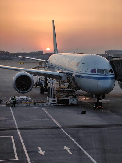 An airplane sits at a gate in the Shunyi District, China. Ground personnel are working around the plane. A baggage carrier loaded with luggage sits adjacent to the plane. The sun is visible in the distance at sunset.