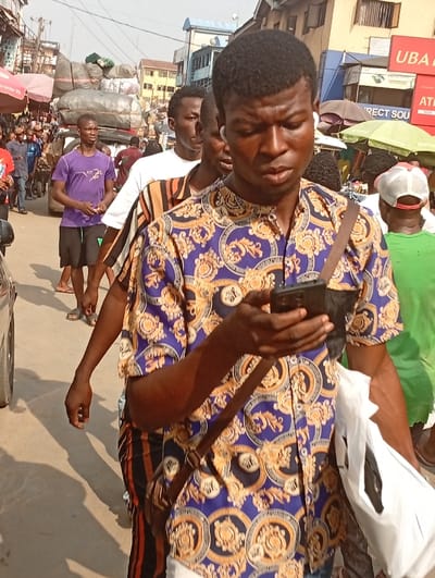 A man in a patterned shirt checks his phone while walking on a busy street. He wears a shoulder bag and carries a white plastic bag. Several other men surround him, and a vehicle laden with goods is visible in the background. Umbrellas and storefronts line the street, creating a bustling market atmosphere.