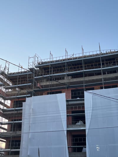 The image depicts a multi-story building under construction, heavily covered in white sheeting, located in Limassol, Cyprus. The building appears to be made of concrete and brick, with scaffolding wrapped around its exterior. Several floors are visible through gaps in the sheeting, revealing unfinished interiors and windows. The sky is clear and blue, suggesting daylight. Thin wires, possibly power lines or communication cables, are strung across the lower part of the image. No people are visible in the scene. The overall impression is of an active construction site in an urban setting.