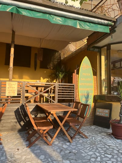 This media file captures an outdoor seating area, likely a cafe or restaurant, with a distinctly beach-themed ambiance. The scene is set on a sunny day, judging by the harsh shadows cast across the stone and wooden patio. A light green surfboard-shaped sign, prominently displaying "PIZZA, SALADS, APETIZERS, SPAGHETTI, DESSERTS, GRILL" and the word "breeze," leans against a yellow-painted wall. In front of it, several wooden folding chairs and tables are arranged on a wooden deck. To the left, a large, tan-colored awning provides shade, with a green tarp hanging from a metal pole.
A smaller, weathered sign with Bulgarian text, "ДЕТСКИ КЪТ" (Children's Corner) and icons of a car and airplane, is mounted on the yellow wall near a large potted plant. Another sign reads "Surf Bus PARKING ONLY VEEDUB ZONE". The overall impression is a casual, relaxed outdoor dining or gathering space, possibly near a beach or coastal area given the surf-inspired decor. The lighting suggests it is daytime, likely late morning or afternoon, with bright sunlight. No people are visible in the image.