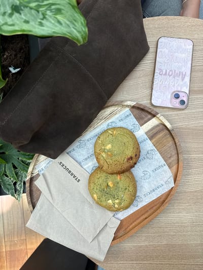 A high-angle shot shows a table setting in what appears to be a Starbucks in Matosinhos, Portugal. A mobile phone with a pink case displaying the word "Amore" sits to the upper left of the frame. A brown suede bag lies partially visible to the left. A wooden plate holds two green cookies, served on paper napkins with Starbucks logos and croissant illustrations. A stack of beige Starbucks napkins is also present on the plate. Green plants are situated below the table's surface, near the bag. The table itself has a light wood grain finish.