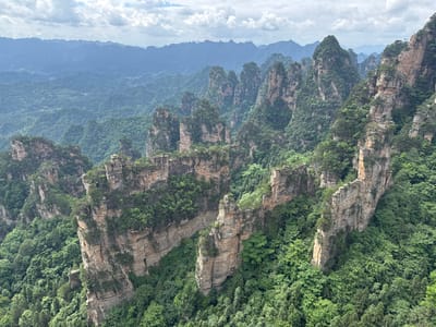 The image depicts a landscape of tall, pillar-like rock formations covered in dense green vegetation. The formations are stratified, with visible layers of rock. In the distance, a hazy blue mountain range is visible. The sky is partially obscured by clouds. This location appears to be in the city of Zhonghu, China. The photograph appears to be rotated 180 degrees.