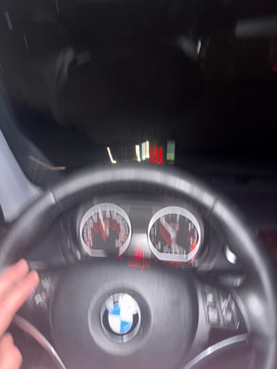 The image shows the interior of a car, specifically a blurred view of the steering wheel and dashboard. The steering wheel has the BMW logo in the center. The dashboard features two circular gauges with red needles. The image is blurry, suggesting motion, possibly taken while the car was in motion. Part of a hand is visible at the bottom right of the image. The left side of the image is dark and indistinct, but appears to be the exterior view through the car window.