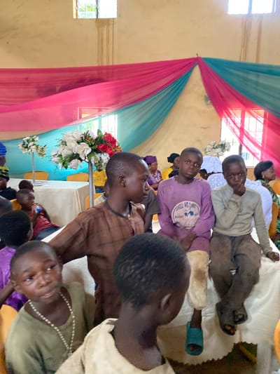 The image depicts an indoor gathering, possibly a community event or celebration, in Riyom, Nigeria. The scene is decorated with draped pink and turquoise fabric, which is suspended from the ceiling, creating a festive backdrop. In the foreground, several children are seated, some looking towards the camera and others engaged in their own activities.

One boy in the center wears a light purple long-sleeved shirt with a graphic that reads "SHELL WE DANCE." To his right, another boy wears a light gray long-sleeved shirt and dark pants, with his hand resting near his chin. To the left of the purple-shirted boy, a child in a brown garment is partially visible. Further left, a young boy in a green shirt with a beaded necklace is looking away. Several other children and adults are present in the background, seated at tables or standing. White floral arrangements are visible on stands, adding to the celebratory atmosphere. Two windows with visible panes are present in the upper part of the image, allowing natural light to enter the space. The overall impression is of a lively, communal event in a modest indoor setting.