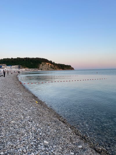 This image captures a serene coastal landscape at what appears to be late afternoon or early evening, in Olginka, Russia.

The scene features a wide, extensive pebble beach in the foreground and mid-ground, composed of various shades of grey, white, and brown stones. The beach gently slopes towards a calm body of water, likely the Black Sea, which occupies the central and right portions of the image. The water near the shore is clear enough to reveal the pebbles beneath, and further out, its surface reflects the soft light of the sky. A distinctive line of evenly spaced red-orange rectangular buoys stretches across the water, marking a designated area, possibly for swimming or boating.

Along the beach, several small figures of people are visible, scattered sparsely. Some individuals appear to be standing or walking, while others might be seated or lounging, enjoying the tranquil environment. Further along the coastline, at the base of the cliff, there are several white structures that resemble beach facilities, such as cabanas or changing rooms.

In the background, on the left side of the image, a lush, green cliff or hillside rises steeply from the water's edge, covered densely with trees and vegetation. The sky above is a clear gradient, transitioning from a pale blue on the left to a soft, warm, almost rosy-white hue towards the horizon on the right, indicative of a peaceful sunset or sunrise.

The overall atmosphere is calm and inviting. There is no visible text in the image.