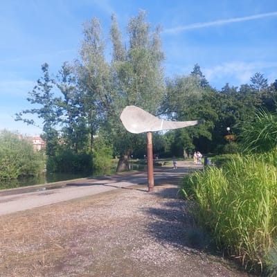 The image depicts a sunny day in a park area in Mrągowo, Poland. In the foreground, a pathway made of stone slabs runs alongside a body of water on the left, bordered by green reeds on the right. A large, abstract sculpture resembling a bird or wing, made of light-colored material, stands on a rusty metal pole in the middle ground. Further back, a paved walking path curves through the park, with several people strolling along it. Trees and dense foliage form a backdrop to the scene, with a glimpse of buildings peeking through the greenery on the left. The sky is clear blue with a few wispy clouds.