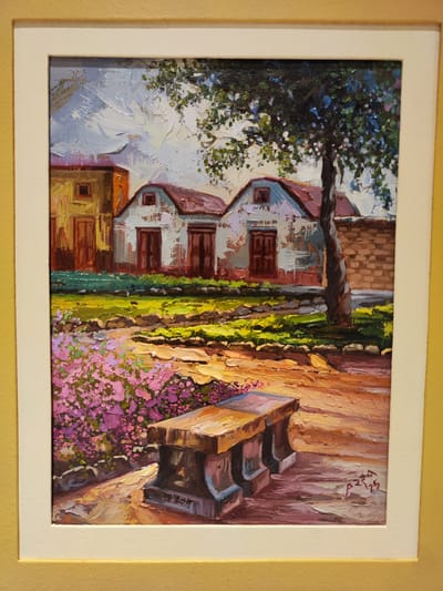 This oil painting, framed in white and mounted on a yellow-gold surface, depicts a vibrant and textured rural or village scene, likely in Tacna, Peru, given the provided context. The artwork is characterized by a heavy impasto technique, where thick layers of paint create visible brushstrokes and texture, particularly in the sky, foliage, and ground.
In the mid-ground, several rustic buildings are visible. Two prominent structures in the center feature reddish-brown gabled roofs and weathered, light-colored walls, possibly white or pale blue, with dark wooden doors or shutters. To the far left, another building, partially obscured, is painted in warm yellow-orange hues. A tall, slender tree with dark brown bark and sparse green foliage stands on the right, casting long shadows across the scene. Behind the tree, a low brick wall extends to the right.
The foreground is dominated by a lush garden or yard area. A patch of bright green grass lies closer to the buildings, while a more rugged, earth-toned pathway or ground area leads towards the viewer. On the left side of the foreground, a large bush bursts with numerous small, vibrant purple and pink flowers, painted with energetic dabs of color. A sturdy, light-colored stone or concrete bench with a flatter, possibly wooden, top is placed in the lower right foreground.
The lighting suggests a warm, sunny day, either late afternoon or early morning, with strong light sources creating distinct shadows and highlights across the landscape. No human figures are present, lending a peaceful and static atmosphere to the scene. The overall impression is one of tranquility and natural beauty.
A signature is visible in the lower right corner of the painting, appearing as "PZ" followed by the numbers "25", likely indicating the artist's initials and the year of creation.