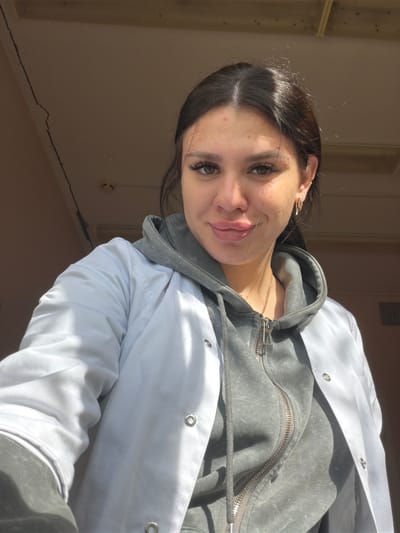 The image is a close-up, selfie-style shot of a young woman with dark hair, light brown eyes, and full lips. She is wearing a white lab coat over a grey hooded sweatshirt. The background appears to be indoors, with a light-colored ceiling and wall, possibly a classroom or laboratory. The lighting suggests it is daytime, with sunlight casting shadows. No specific location in the city of Гаревая, Russia, can be determined from the image.