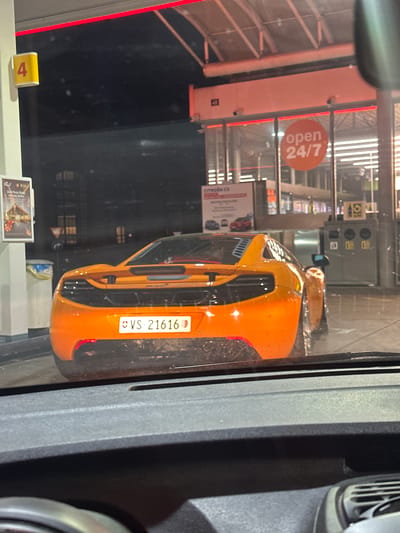 The image shows an orange sports car, likely a McLaren, with the Swiss license plate VS-21616. The car is parked at a gas station, visible through a window. The sign "open 24/7" is visible inside the gas station. A sign displays "CITROEN C5". A gas pump marked with a yellow "4" is also present. The location appears to be a gas station in Winterthur, Switzerland.