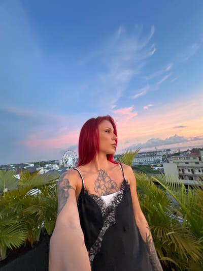 A woman with red hair, tattoos on her chest and arms, is standing in what appears to be Tibubeneng, Indonesia, based on the presence of a Ferris wheel in the background and the city landscape. The photo is taken at a low angle, showing a partly cloudy sky with colors of sunset. She is wearing a black garment with a white lace. Green tropical plants surround her. Buildings are visible in the background.