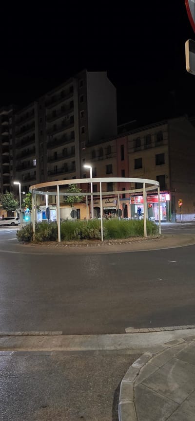 The image depicts a night scene in Huesca, Spain, featuring a roundabout with a circular white structure in its center. The structure is adorned with greenery and appears to be a public art installation or a decorative element. Surrounding the roundabout are multi-story buildings, some of which are illuminated and house businesses. One of these businesses is clearly identified as "DONER KEBAB" with a red neon sign. Streetlights cast a glow on the scene, illuminating the road and the surrounding architecture. A white car is visible on the left side of the image, parked near a traffic light. The overall impression is that of a typical urban environment at night, with commercial activity and residential buildings coexisting.